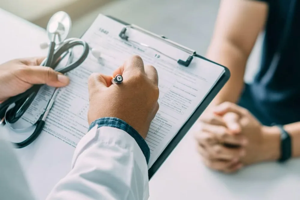 Médico preenchendo a ficha de avaliação em uma prancheta, com estetoscópio sobre a mesa, durante consulta em consultório