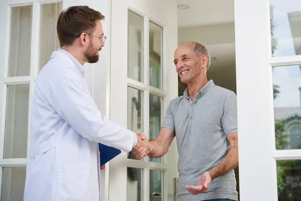Médico de jaleco branco cumprimentando um paciente idoso na porta de casa, em atendimento domiciliar